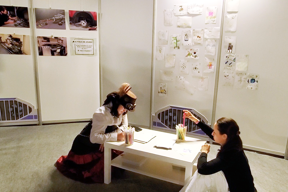 Deux adultes, dont l'une en style streampunk, dessinant agenouillées à une table basse. Des dessins sont affichés au mur derrière.