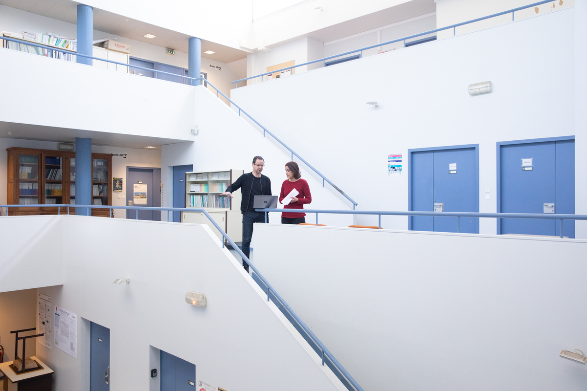 Un chercheur et une chercheuse au milieu des couloirs et escaliers d'un bâtiment